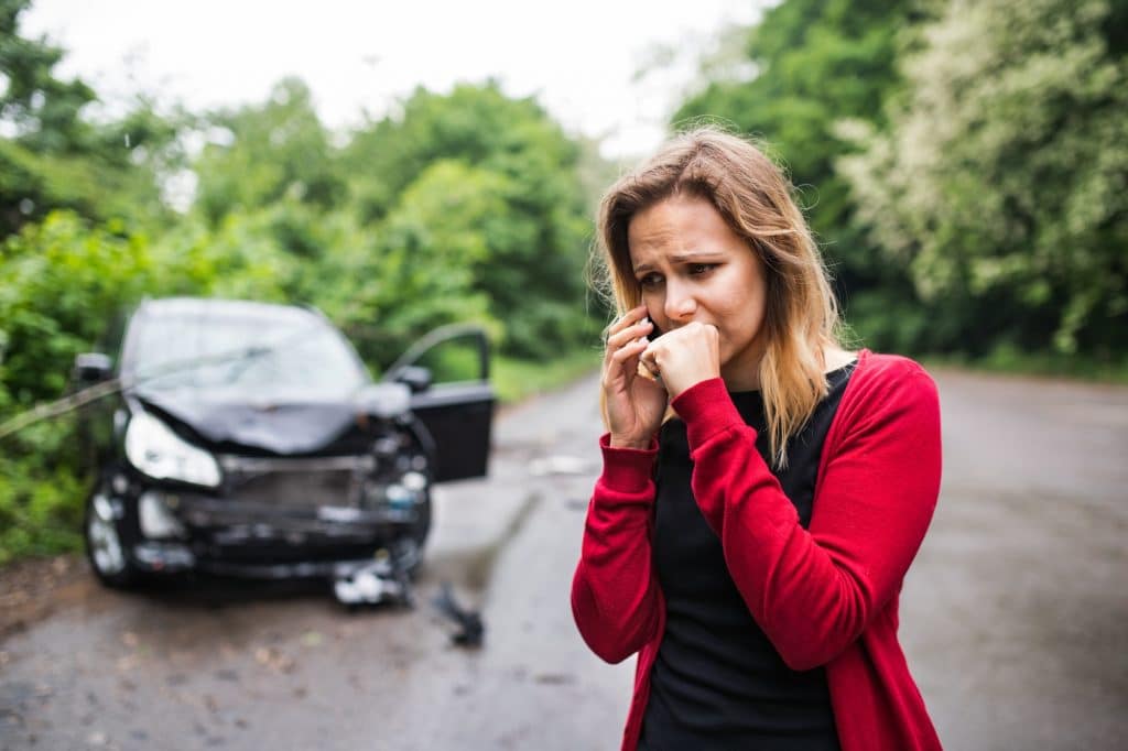 Una giovane donna con lo smartphone dall'auto danneggiata dopo un incidente d'auto, facendo una telefonata.